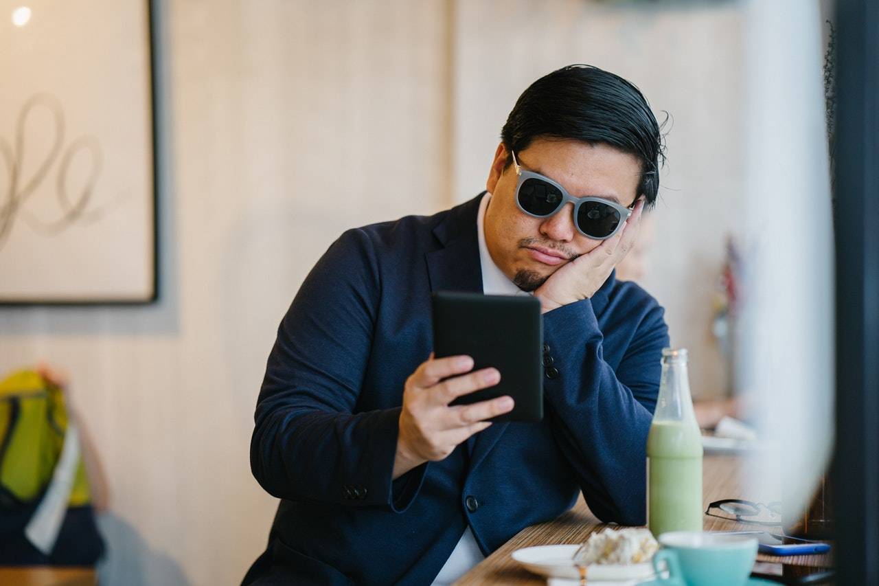 Man with sunglasses in a cafe looking bored whilst he looks at his phone 