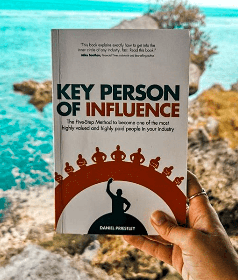 Hand holding book Key Person of Influence by Daniel Priestly in front of a seashore
